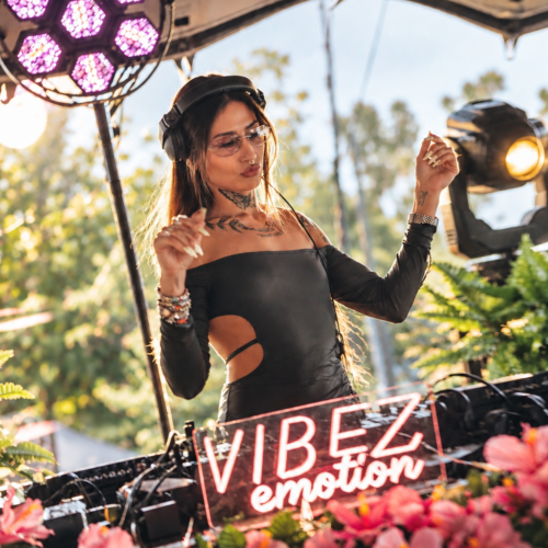 A DJ at the decks behind a neon Vibez Emotion sign, outdoors among flowers