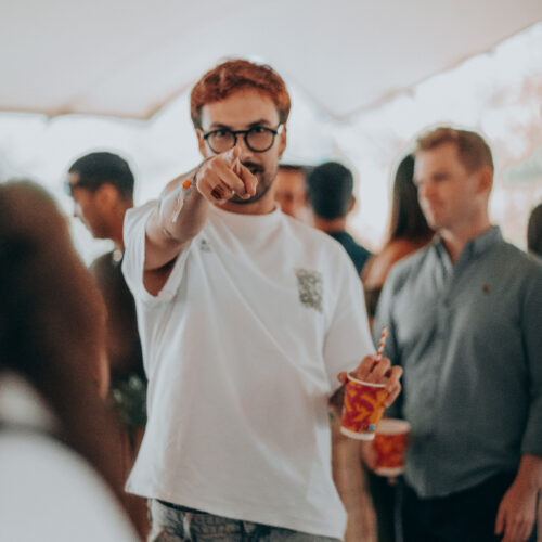 A man in glasses pointing at the camera, drink in hand