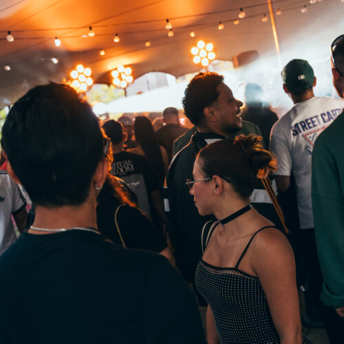 A crowd under a tent with string lights, facing the stage